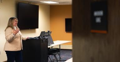 Dr. 珍·博萨德在商学院的技术教室前面讲话, 位于林肯的弗雷德·布朗中心. 一扇打开的门部分遮住了图像, 博萨尔身后是两个大电视屏幕, 一个平板电脑和一个科技柜.